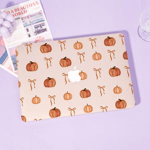 May include: A light beige laptop hard shell case featuring a repeating pattern of orange pumpkins and brown ribbon bows. The center of the case displays a white Apple logo cutout. The design is printed across the entire surface of the cover.