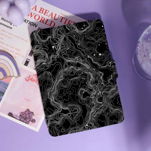 May include: Black and white topographic map design on a rectangular case. The case has a flap closure. The words "A BEAUTIFUL WORLD" are visible on a nearby card. The case is on a purple surface.