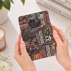 May include: A rectangular card featuring a colorful illustration of three shelves filled with books, pumpkins, a bubbling cauldron, a sleeping black cat, and a skull planter. The design includes celestial symbols and the word Spells on a book spine.