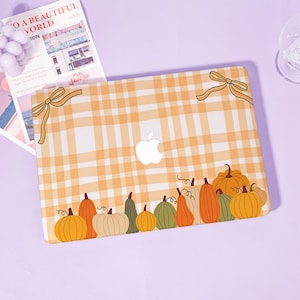 Può includere: Una custodia rigida per laptop con un motivo a quadri arancione chiaro e bianco. Il design include due fiocchi color marrone chiaro negli angoli superiori e una fila di varie zucche in tonalità arancione, verde e crema lungo il bordo inferiore.