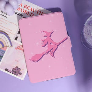 May include: A pink tablet case featuring a purple silhouette of a witch on a broomstick, surrounded by star shapes. The case has a magnetic closure. The text "A BEAUTIFUL WORLD" is visible in the background.