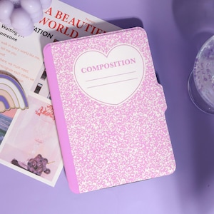 May include: A pink composition notebook with a heart-shaped design and the word "COMPOSITION" in the center. The notebook has a speckled pattern and a pink spine. The words "A BEAUTIFUL WORLD" are visible in the background.
