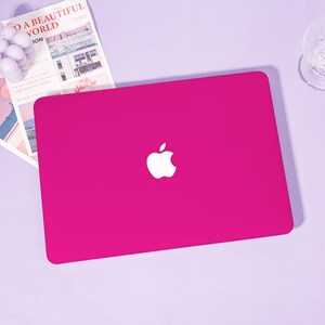 May include: A vibrant magenta laptop with a white Apple logo. The laptop is on a light purple surface, with a magazine and a glass in the background. The magazine has the text "TO A BEAUTIFUL WORLD".