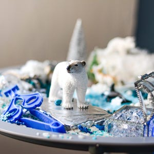 Op de afbeelding: Een wit ijsberenbeeldje dat op een zilveren platform staat, omringd door blauw en wit plastic speelgoed en een doorzichtige glazen doos met zilveren accenten. Het speelgoed ligt op een zilveren dienblad.