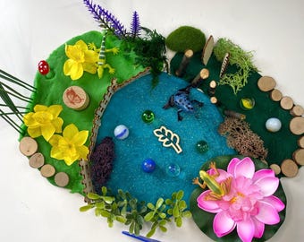 Puede incluir: Una escena de juego sensorial con un estanque azul, colinas verdes y una flor de loto rosa. La escena incluye ranas de juguete, canicas y detalles de madera. Unas pinzas azules están en primer plano. Diseñado para el juego de los niños.