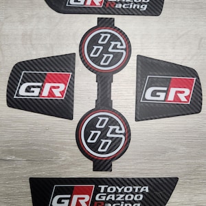 Może przedstawiać: Zestaw czarnych naklejek o fakturze włókna węglowego z logo "GR" w kolorze białym i czerwonym oraz napisem "TOYOTA GAZOO Racing". Zawiera okrągłe i prostokątne kształty, idealne do personalizacji samochodów.