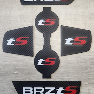 May include: A set of black carbon fiber automotive accessories. The pieces feature white and red text that reads "BRZtS" and "tS". The accessories are arranged on a light gray surface.