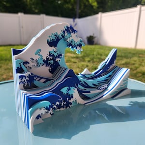 Pode incluir: Uma escultura em camadas 3D azul e branca de uma onda, inspirada na famosa gravura japonesa em madeira "A Grande Onda de Kanagawa" de Hokusai.