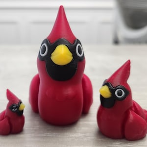 May include: Three red cardinal figurines with black and yellow details. The figurines vary in size, with one large and two smaller versions. The birds have a distinctive crest and are posed in a resting position.