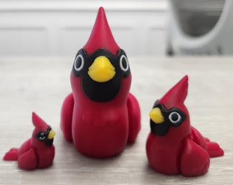 Può includere: Tre statuette di cardinali rossi con dettagli neri e gialli. Le statuette variano in dimensioni, con una grande e due più piccole. Gli uccelli hanno una cresta distintiva e sono in posizione di riposo.