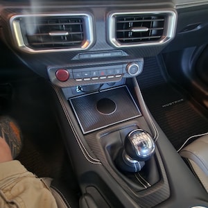 Pode incluir: Interior de um carro com painel e consola. Inclui saídas de ar, um botão de arranque vermelho e uma alavanca de velocidades. A consola tem um padrão de fibra de carbono e costuras brancas. O tapete do carro diz "MUSTANG".