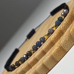 May include: A beaded bracelet with blue and gold beads. The bracelet is presented on a bamboo stand with a black cord. The small, round beads have varied colours and textures. The bracelet is a piece of jewellery.