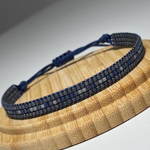 May include: A blue and silver beaded bracelet with a sliding knot closure. The bracelet is displayed on a wooden surface.