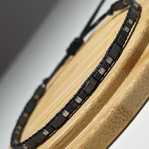 May include: A black and silver beaded bracelet with a black cord. The bracelet is on a wooden surface.
