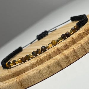 Könnte beinhalten: Ein braunes und goldenes Perlenarmband mit einem schwarzen Band. Das Armband liegt auf einer Holzoberfläche.