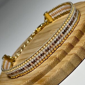 Könnte beinhalten: Ein goldenes Armband mit weißen und braunen Perlen. Das Armband liegt auf einer Holzoberfläche.
