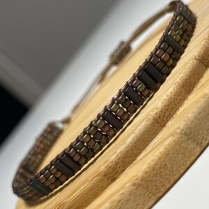 May include: A brown and gold beaded bracelet with a dark brown leather cord. The bracelet is on a wooden surface.