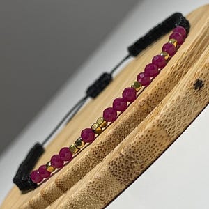 May include: A pink and gold beaded bracelet with a black cord. The bracelet is tied with a knot and is on a wooden surface.
