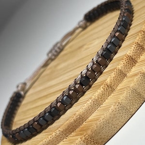 May include: A brown, black and grey beaded bracelet with a braided cord fastening. The bracelet is displayed on a wooden surface.
