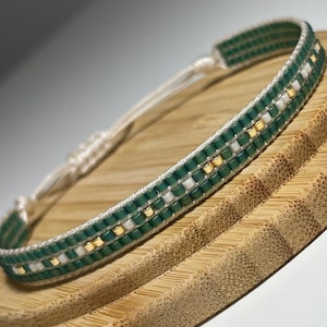 May include: A green and white beaded bracelet with gold accents. The bracelet is on a wooden surface.
