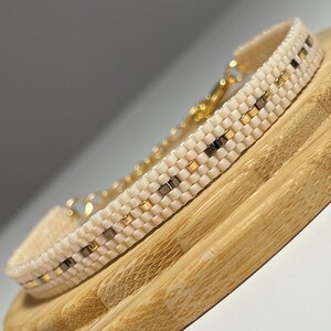 Könnte beinhalten: Ein zartes Armband aus weißen Perlen mit goldenen Akzenten. Das Armband besteht aus kleinen quadratischen Perlen und hat ein schlichtes, minimalistisches Design.