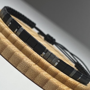 May include: A black and silver beaded bracelet with a black cord. The bracelet is on a wooden surface.