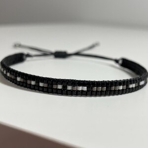 May include: A black beaded bracelet with white and grey beads. The bracelet has a black cord with a sliding knot for adjusting the size.