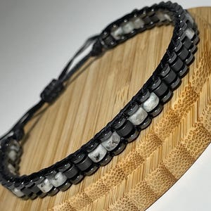 May include: Black and white beaded bracelet with a sliding knot closure. The bracelet features alternating black and white stone beads, creating a striking contrast. The bracelet is displayed on a round, light brown wooden surface.