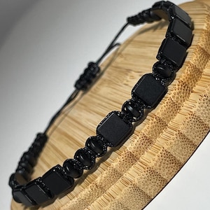 Könnte beinhalten: Schwarzes Perlenarmband mit quadratischen und runden Perlen. Das Armband ist mit einem Schiebeknotenverschluss verstellbar. Die Perlen sind auf eine schwarze Kordel gefädelt. Das Armband wird auf einem Holzständer präsentiert.