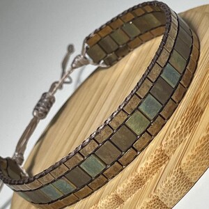 Può includere: Un braccialetto fatto a mano con un design a mosaico. Il braccialetto presenta piccole tessere quadrate nelle tonalità del marrone, del verde e dell'oro, disposte in un motivo ripetuto. Il braccialetto è regolabile con un cordino intrecciato.