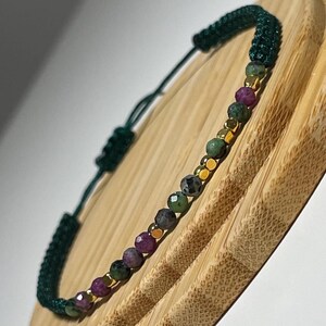 Può includere: Un braccialetto in cordoncino verde con una fila di piccole perline di pietre preziose multicolori. Le perline sono un mix di verde, rosa e nero.