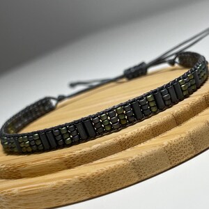 Peut inclure: Un bracelet en perles noires et vertes avec une fermeture en cordon noir. Le bracelet est posé sur une surface en bois.