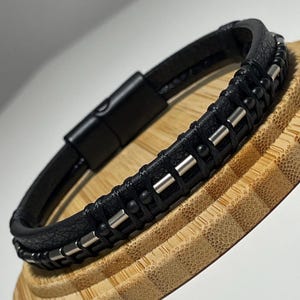 Könnte beinhalten: Schwarzes Lederarmband mit silberfarbenen Akzenten und einem Magnetverschluss. Das Armband hat ein geflochtenes Design mit kleinen, runden Perlen und rechteckigen Metallstäben. Es wird auf einem Holzständer präsentiert.