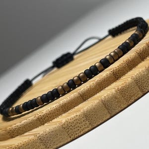 May include: A black and gold beaded bracelet with a black cord. The bracelet is tied with a knot and is lying on a wooden surface.