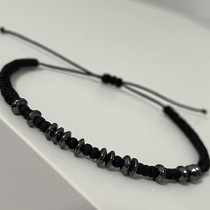 May include: Black adjustable bracelet featuring a series of dark gray metal beads and black cord. The bracelet has a sliding knot closure for a customizable fit. The design is simple and modern, suitable for everyday wear.