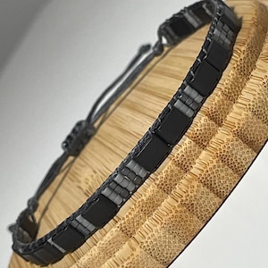 May include: A black and grey beaded bracelet with a sliding knot fastening. The bracelet features rectangular black beads with smaller grey beads. It is displayed on a textured wooden surface.