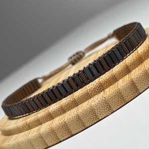 May include: A brown and black beaded bracelet with a braided cord fastening. The bracelet is on a wooden surface.