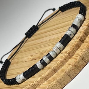 May include: Black and white beaded bracelet with a macrame closure. The bracelet features alternating black and speckled white beads. The bracelet is displayed on a wooden tray.