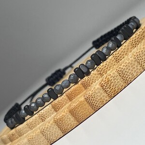 Könnte beinhalten: Ein schwarzes Perlenarmband mit Makramee-Verschluss. Das Armband hat eine Kombination aus dunkelgrauen, facettierten Perlen und schwarzen, rechteckigen Perlen. Das Armband wird auf einer strukturierten Holzoberfläche präsentiert.