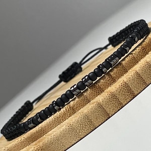 May include: Black beaded bracelet with silver-tone metal accents. The bracelet has a woven adjustable closure. The bracelet is displayed on a wooden surface.
