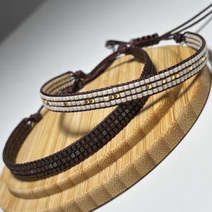 May include: Two beaded bracelets, one brown and one white, with gold accents. The bracelets are displayed on a wooden surface.