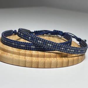 May include: Two beaded bracelets, one dark blue and one silver, on a wooden surface.