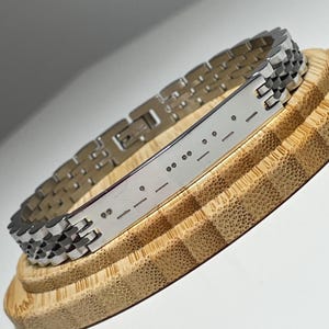 Könnte beinhalten: Ein silberfarbenes Metallarmband mit einer rechteckigen Platte mit Morsecode. Das Armband hat ein Kettenglied-Design und wird auf einem hölzernen, mehrstufigen Ständer präsentiert. Die Morsecode-Platte hat eine Reihe von Punkten und Strichen.