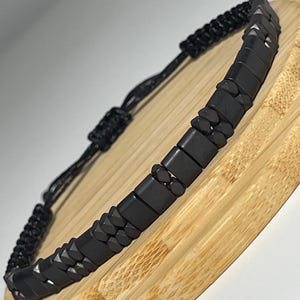 Könnte beinhalten: Schwarzes Perlenarmband mit geflochtenem schwarzem Kordel. Das Armband hat quadratische und abgerundete Perlen. Das Armband wird auf einer hellbraunen Holzoberfläche präsentiert.