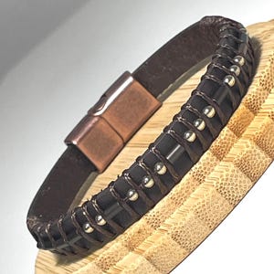 Könnte beinhalten: Ein braunes Lederarmband mit Magnetverschluss. Das Armband hat einen dunkelbraunen, gewebten Abschnitt mit silberfarbenen Metallnieten. Der Verschluss ist kupferfarben. Das Armband wird auf einer Holzoberfläche präsentiert.