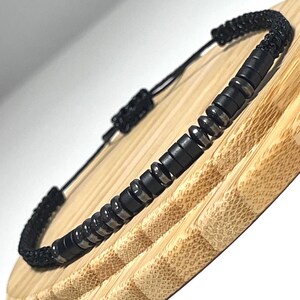 Könnte beinhalten: Schwarzes Perlenarmband mit geflochtenem schwarzem Kordel. Das Armband hat eine Reihe von schwarzen und silberfarbenen zylindrischen Perlen. Das verstellbare Kordel ermöglicht eine bequeme Passform. Das Armband wird auf einer hellbraunen Holzoberfläche präsentiert.