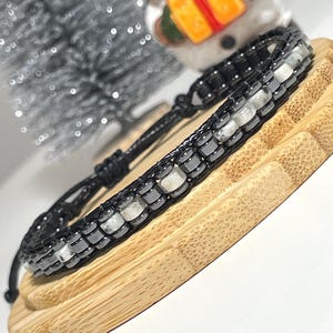 May include: A black wrap bracelet with alternating black and white beads. The bracelet is made of a woven material and has a sliding knot closure. The bracelet is displayed on a wooden surface.