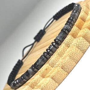 May include: A black and grey beaded bracelet with an adjustable sliding knot. The bracelet features small, round, grey beads with black accents. The adjustable cord is black. The bracelet is displayed on a light brown, textured surface.