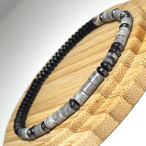 May include: A beaded bracelet featuring a mix of black, gray, and marbled gray beads. The bracelet is made of small, round black beads, interspersed with gray and marbled gray rectangular beads and black cylindrical beads. The bracelet is displayed on a wooden surface.