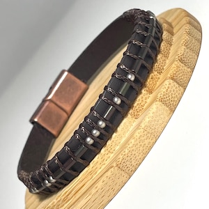 May include: A brown leather bracelet with a woven center section. The center features dark brown bars, silver beads, and brown stitching. The bracelet has a copper-colored magnetic clasp.